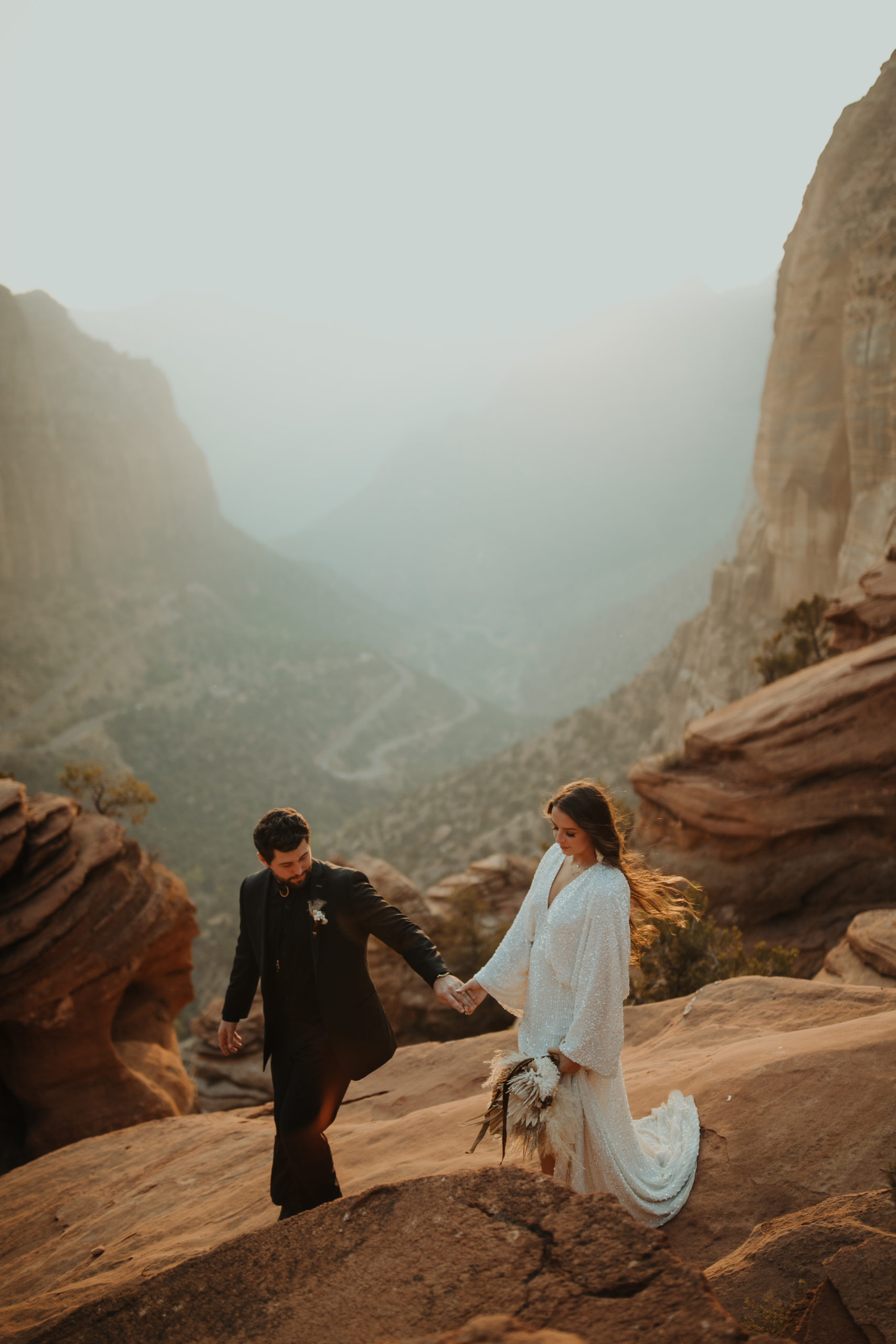 Desert boho couple walking away from cliff with sunset behind them