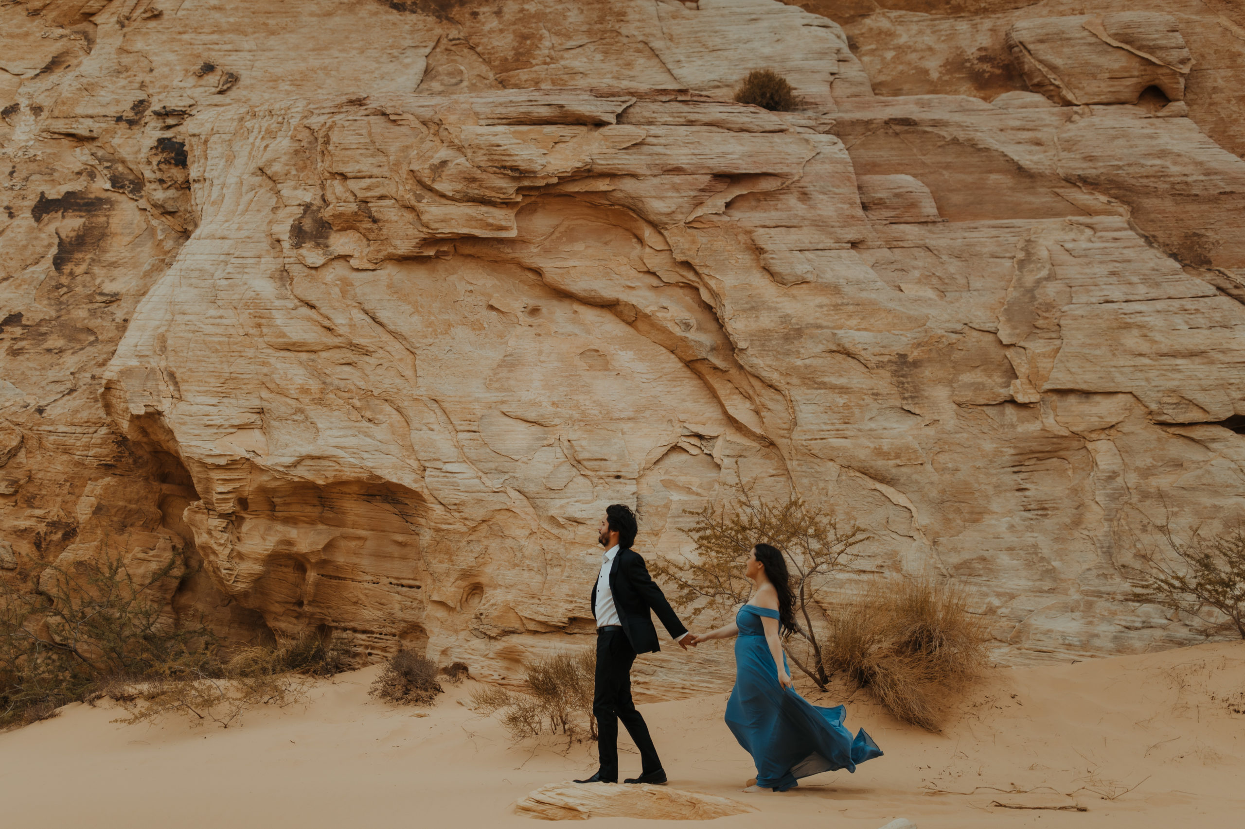 Couple in formal attire running with tan rocks behind them