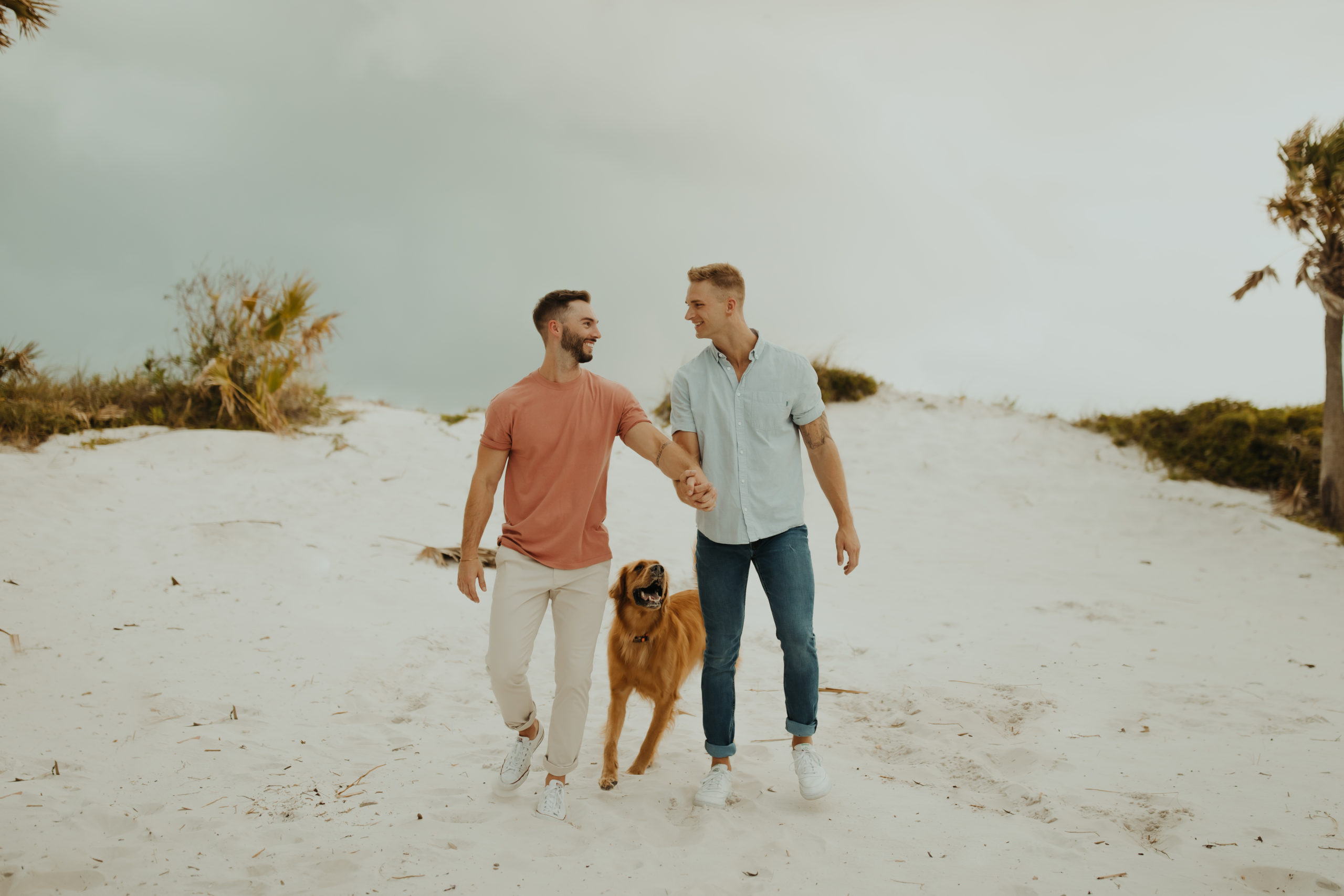 Two men holding hands wearing pastel colors with their golden retriever in between them at a beach in Florida