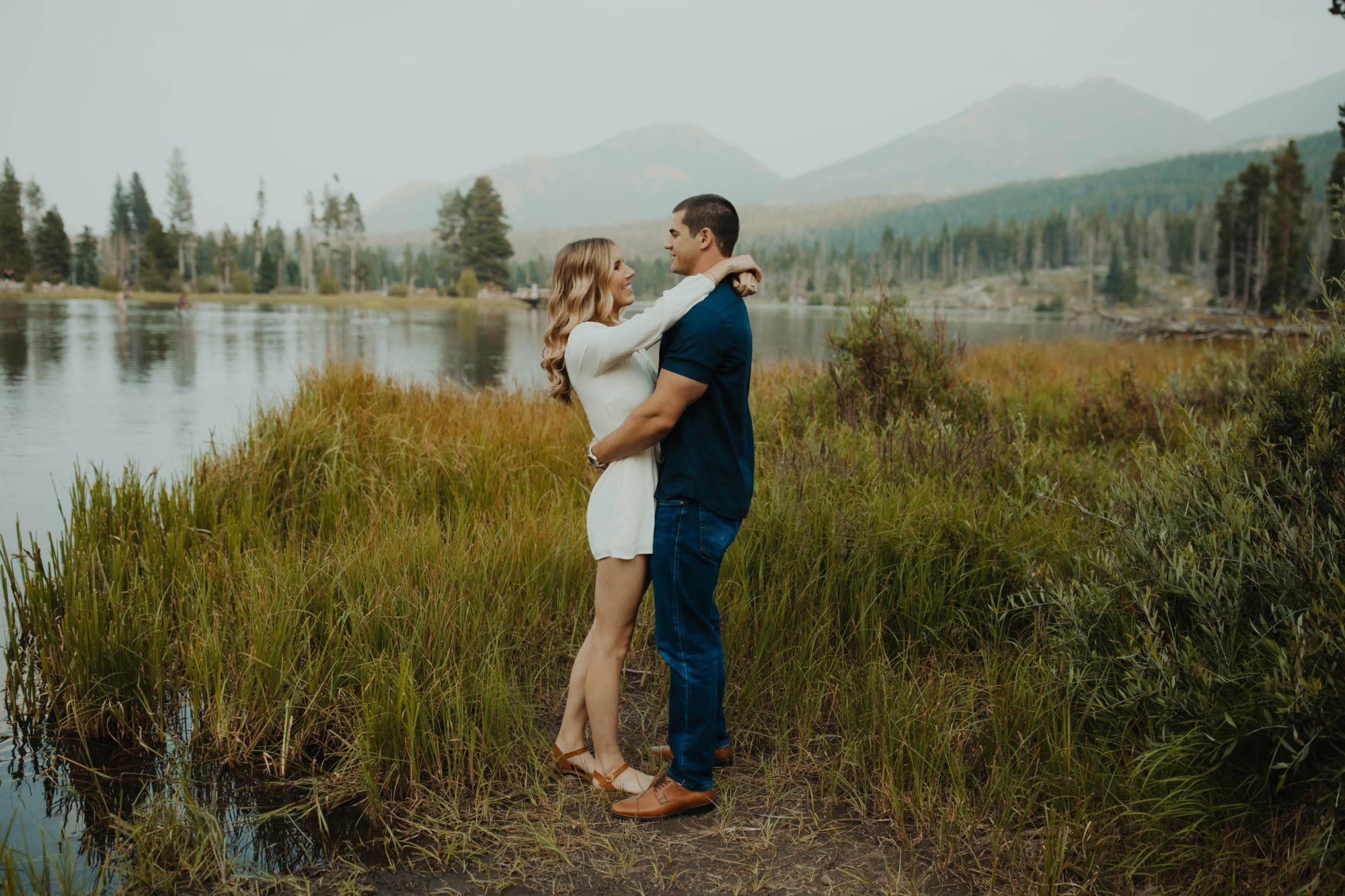 Fiancee wrapping her arms around her boyfriend in front of a lake in the PNW with mountains silhouetted in the  background