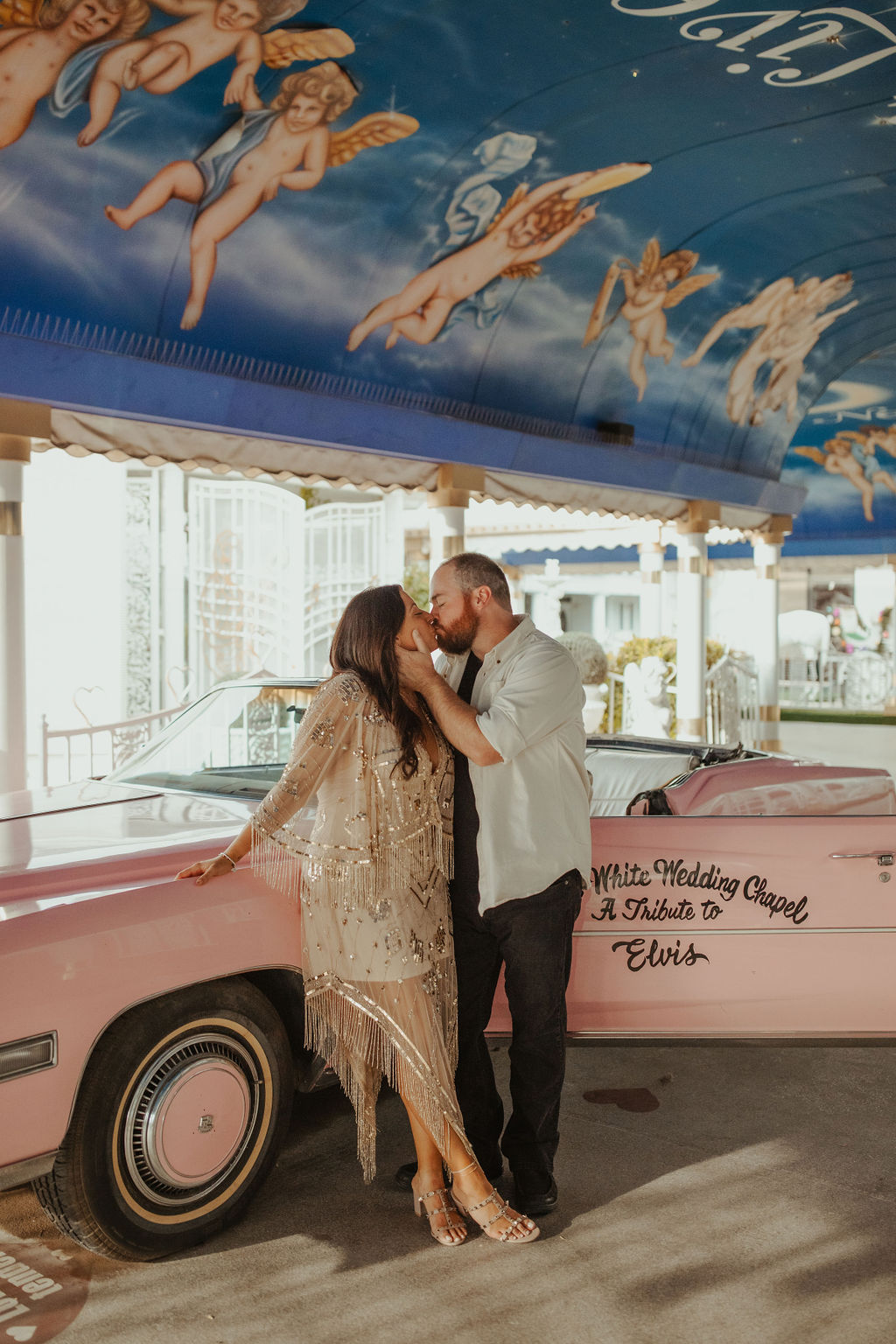 Husband and Wife standing in front of the little white chapel's pink convertible car while kissing