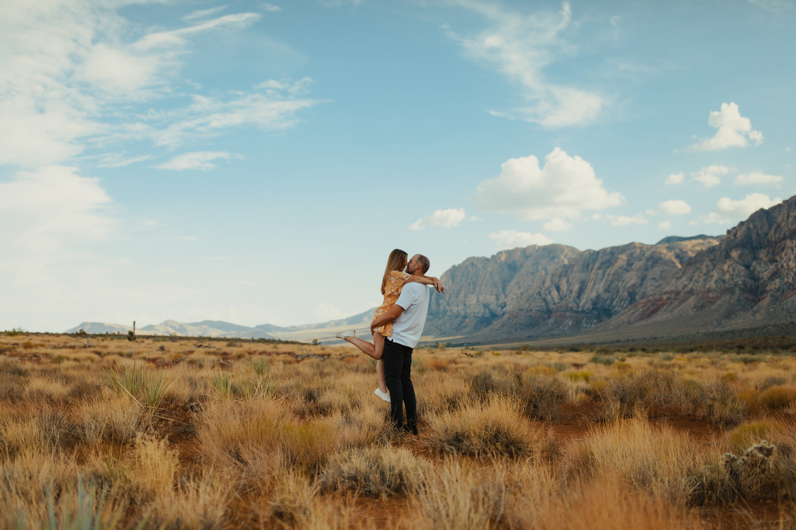 boyfriend picking girlfriend up in a field in the desert with mountains in the background during their engagement session