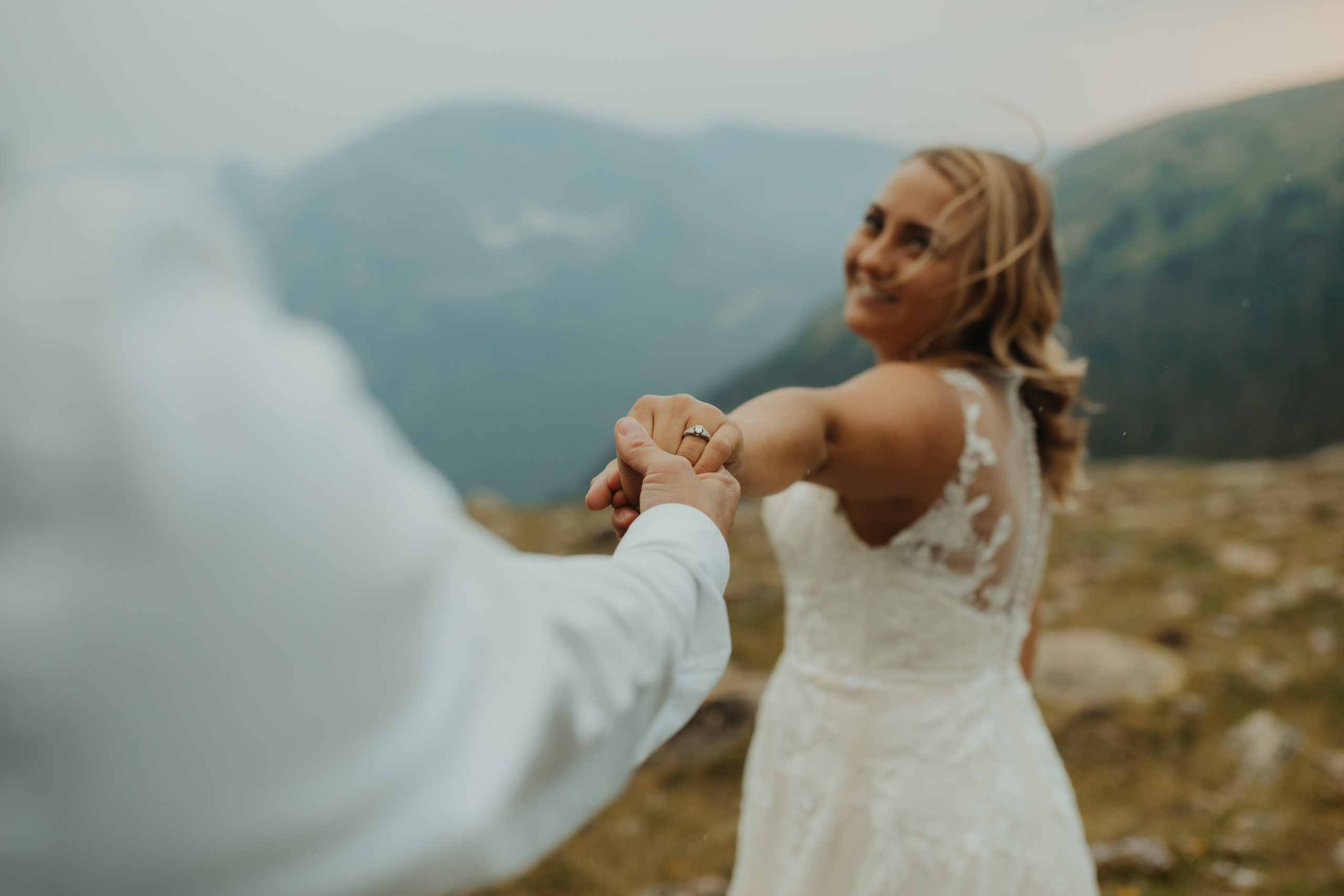 bride holding grooms hands with her and the rocky mountains blurred in the backdrop with the focus point being her engagement ring