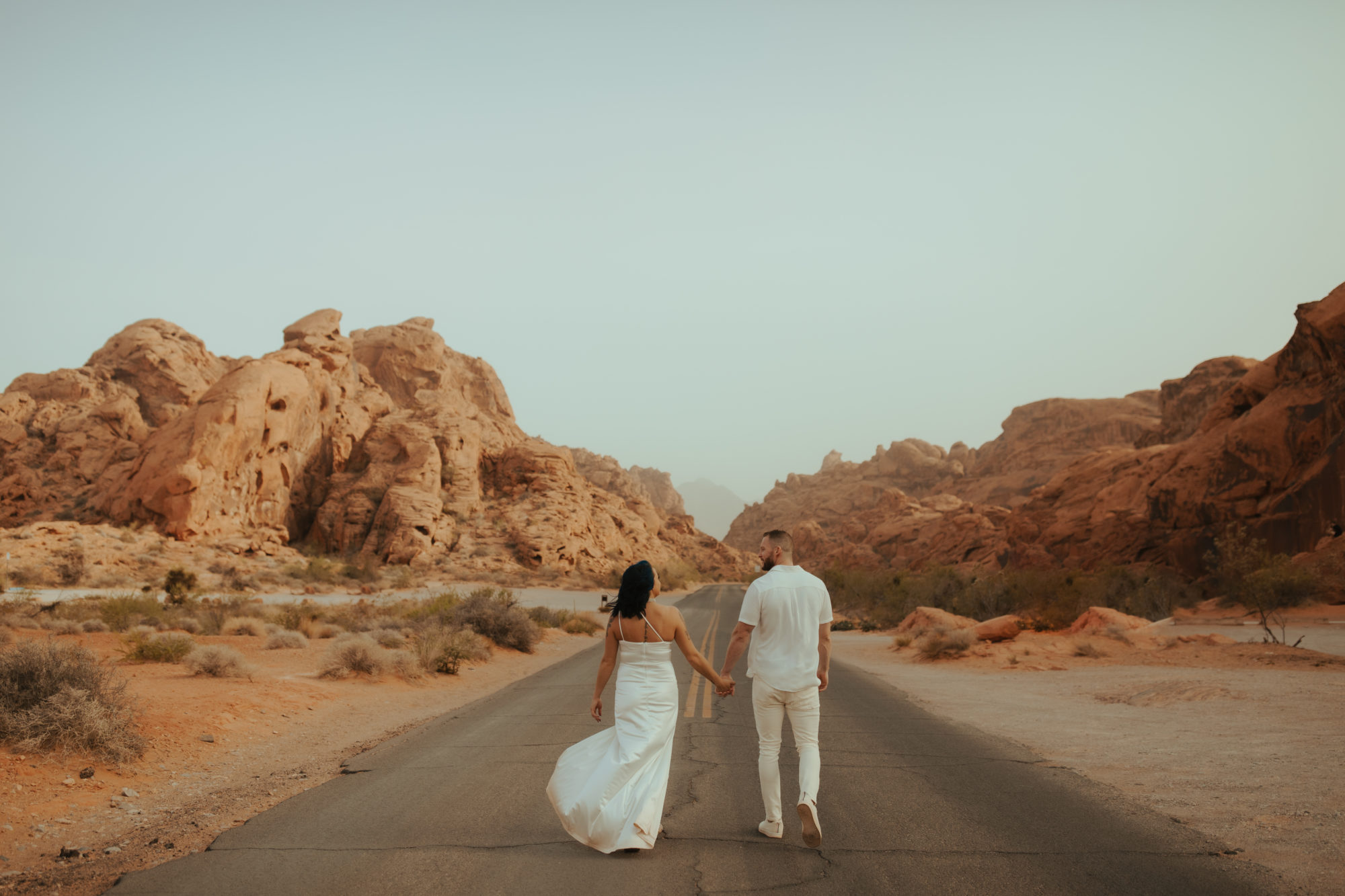 engaged couple walking away from the camera holding hands down a road in valley of fire during a dust storm