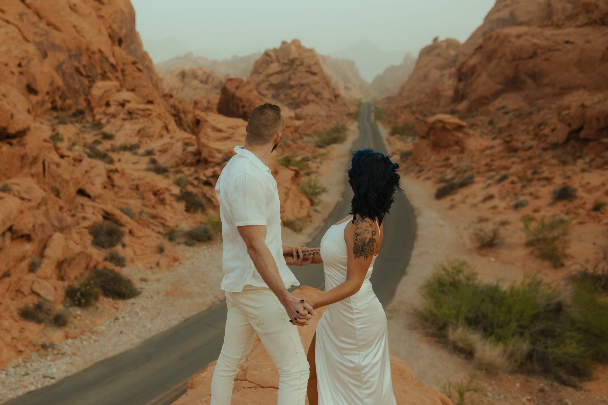 engaged couple holding hands with their arms outstretched while standing on a cliff in valley of fire that overlooks the road