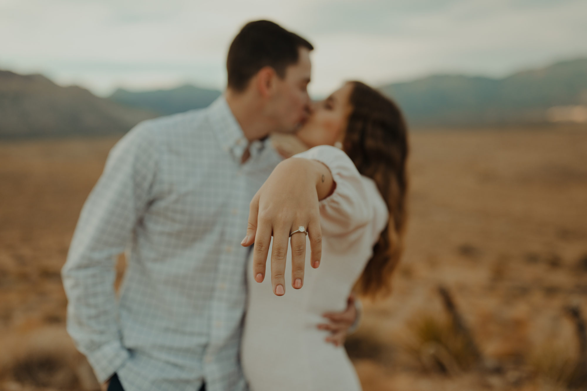 upclose of engagement ring and couple kissing blurred in the background in the desert