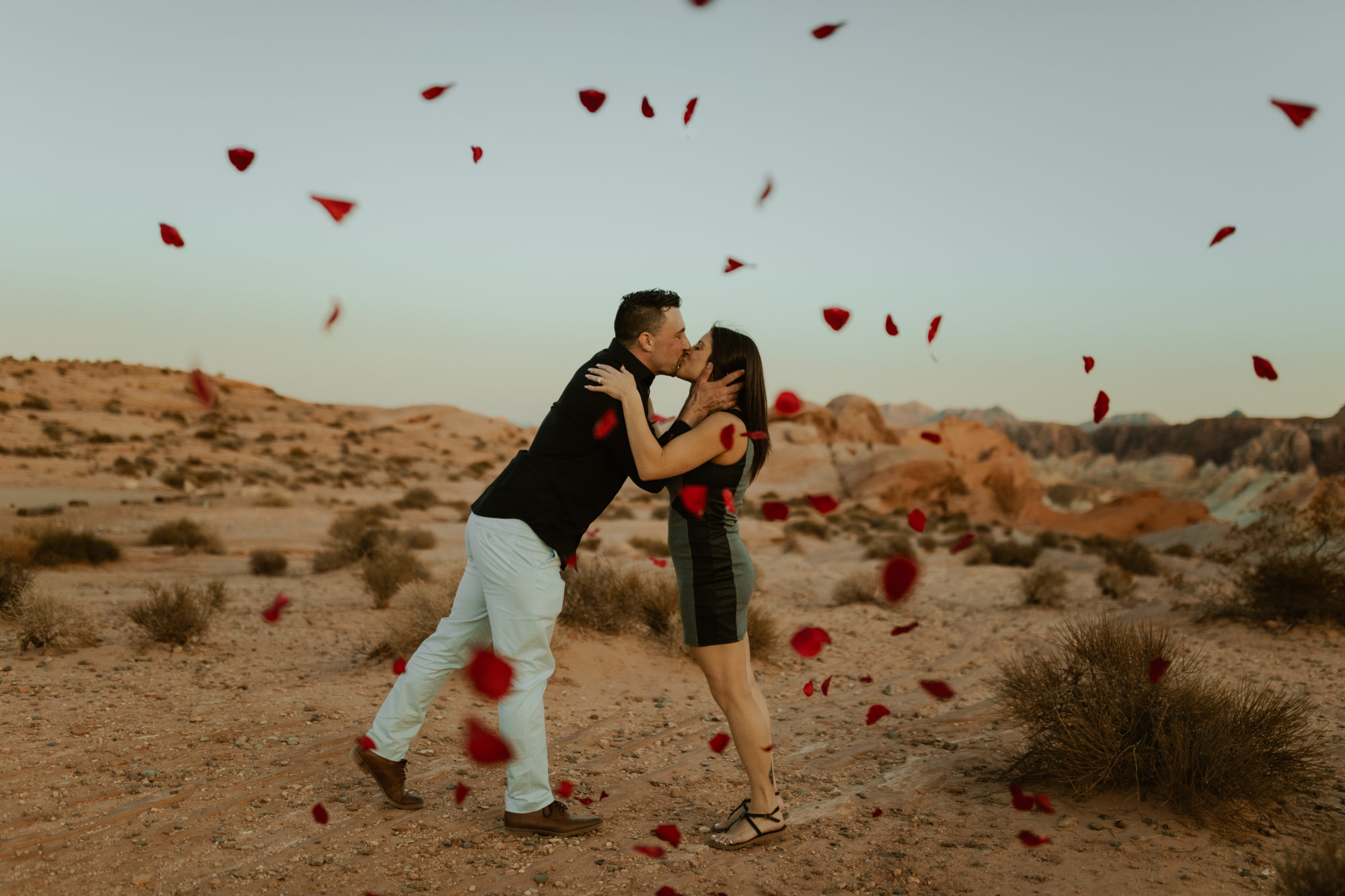 newly engaged couple kissing while rose petals are being thrown at them in the desert after he just proposed to his girlfriend