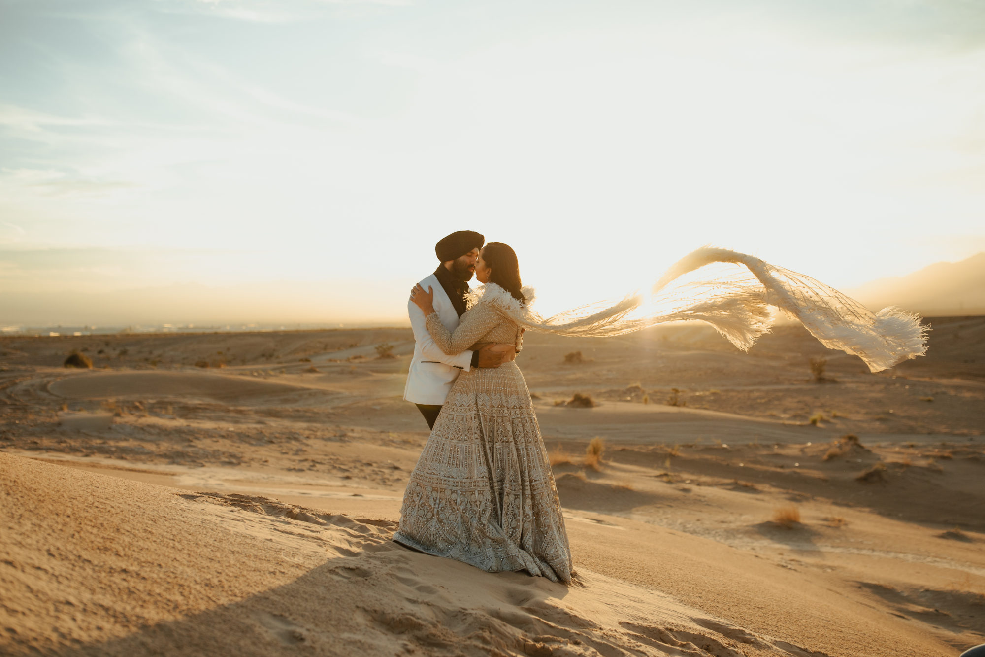 bride and groom standing in the sand dunes of the nevada desert during golden hour as her cape blows in the wind and the groom leans in for a kiss